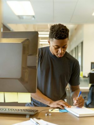 A young man taking notes next to a desktop computer